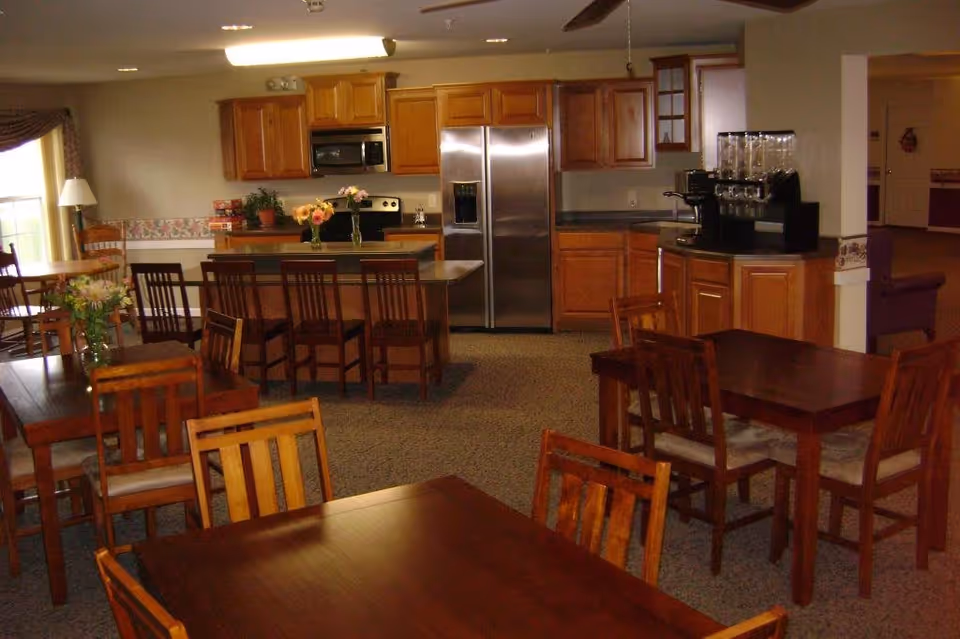 A communal dining area with multiple wooden tables and chairs arranged around the room. In the background, there is a kitchen area with wooden cabinets, a stainless steel refrigerator, microwave, stove, and a coffee machine on the counter. The room is well-lit with natural light coming from a window on the left side.