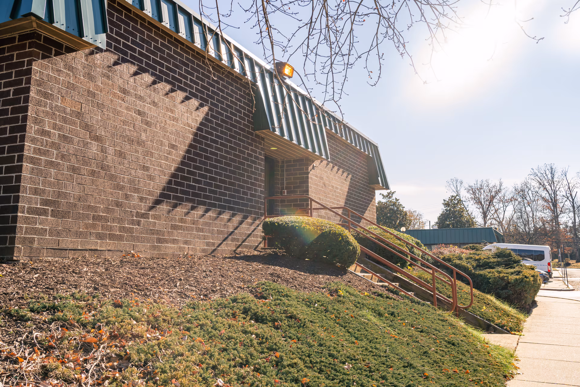 Exterior view of a brick building with a green metal awning and a ramp with handrails leading to an entrance. There are bushes and landscaping along the side of the building, and a white van is parked on the driveway. Trees without leaves are visible in the background under a clear sky.