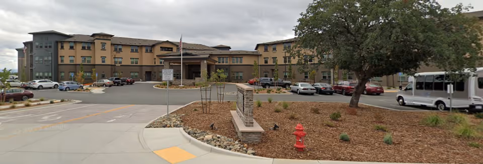 Front exterior of a multi-story senior living building with a circular driveway, parked cars, and landscaped grounds.