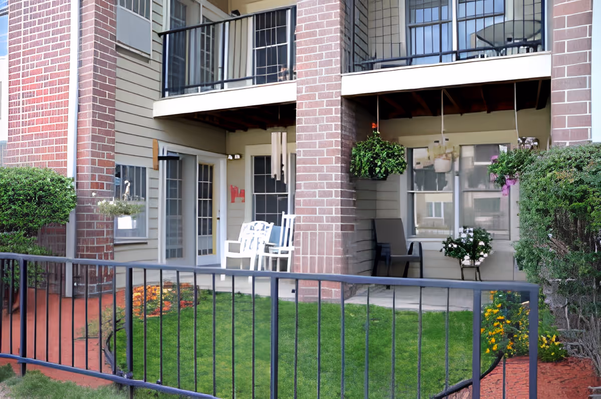 Outdoor patio area of a senior living facility with two white chairs, hanging flower pots, green grass, and a black metal fence in front. The building has brick pillars and beige siding with windows and glass doors.