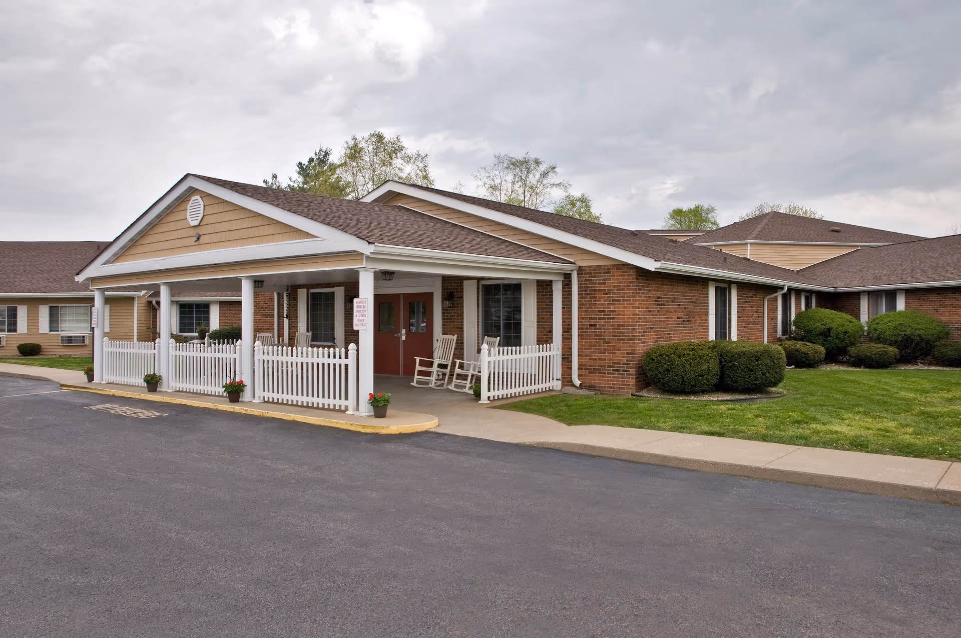 Exterior view of Mitchell Manor senior living facility showing a single-story brick building with a covered entrance supported by white columns and a white picket fence. There are two rocking chairs on the porch, small potted plants, trimmed bushes, and a paved driveway in front. The sky is cloudy.