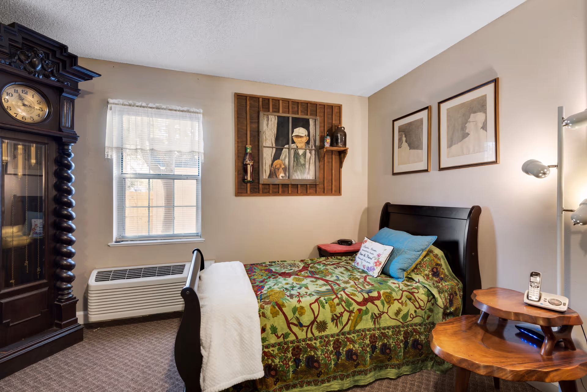 A cozy bedroom in a retirement center featuring a dark wooden bed with a colorful floral bedspread and pillows. There is a wooden grandfather clock on the left side, a window with sheer curtains, and framed artwork on the walls. A wooden side table holds a cordless phone and a lamp with multiple adjustable lights.