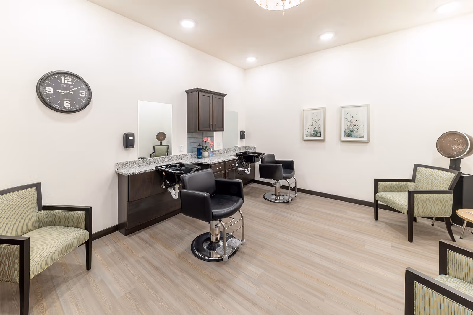 Bright salon room with two styling chairs and wash sinks, several waiting armchairs, a wall clock and framed art.
