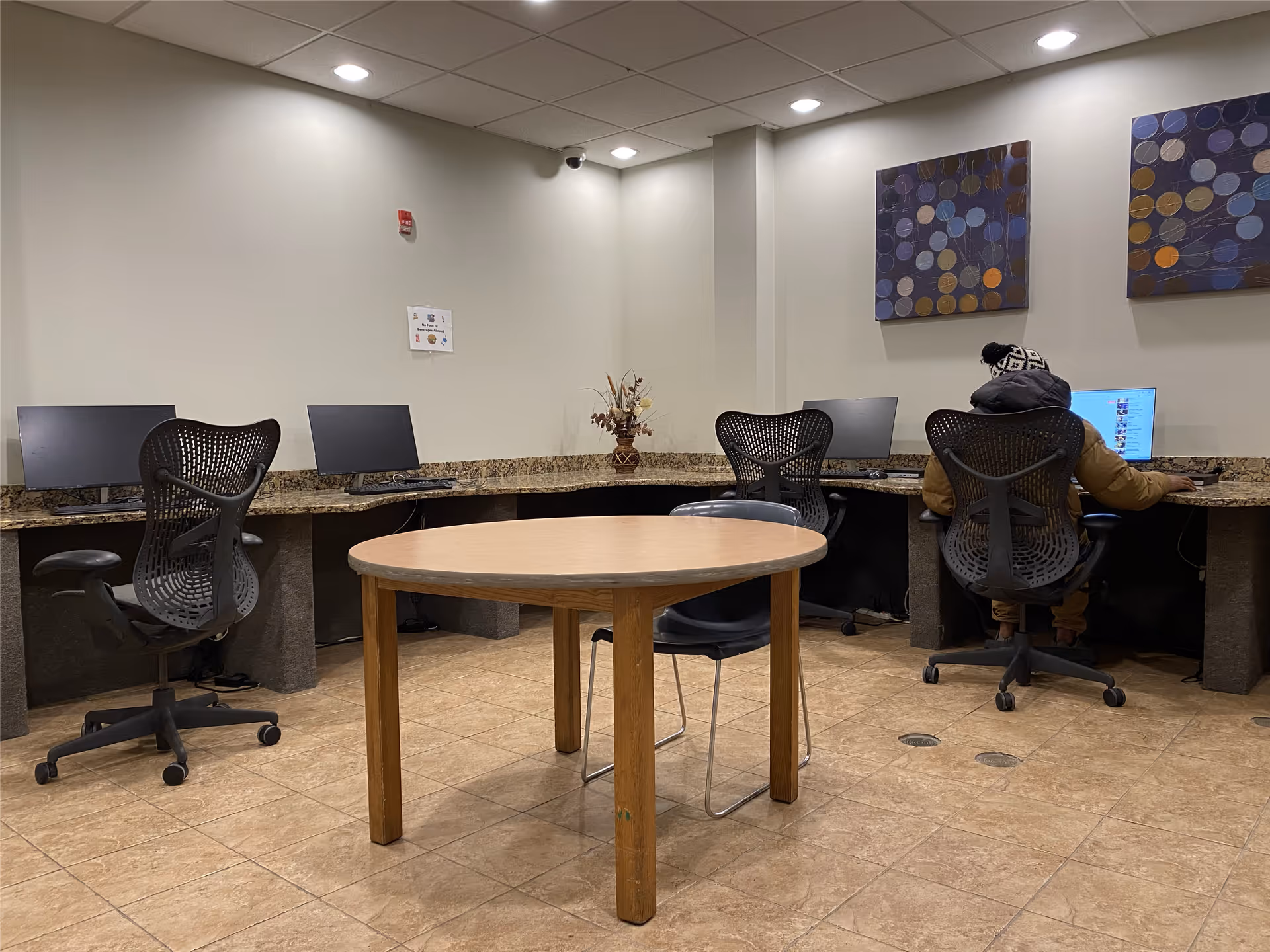 A computer room with four black office chairs and four desktop computers on a granite countertop along the walls. A person wearing a winter jacket and hat is seated at one of the computers. In the center of the room is a round wooden table with two black chairs. The walls are light-colored with two abstract paintings hanging on the right side.