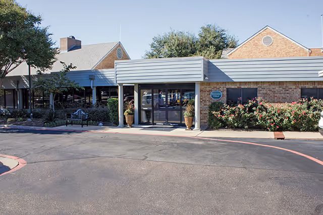 Front entrance of a single-story brick senior living building with an awning, potted plants, and a driveway.