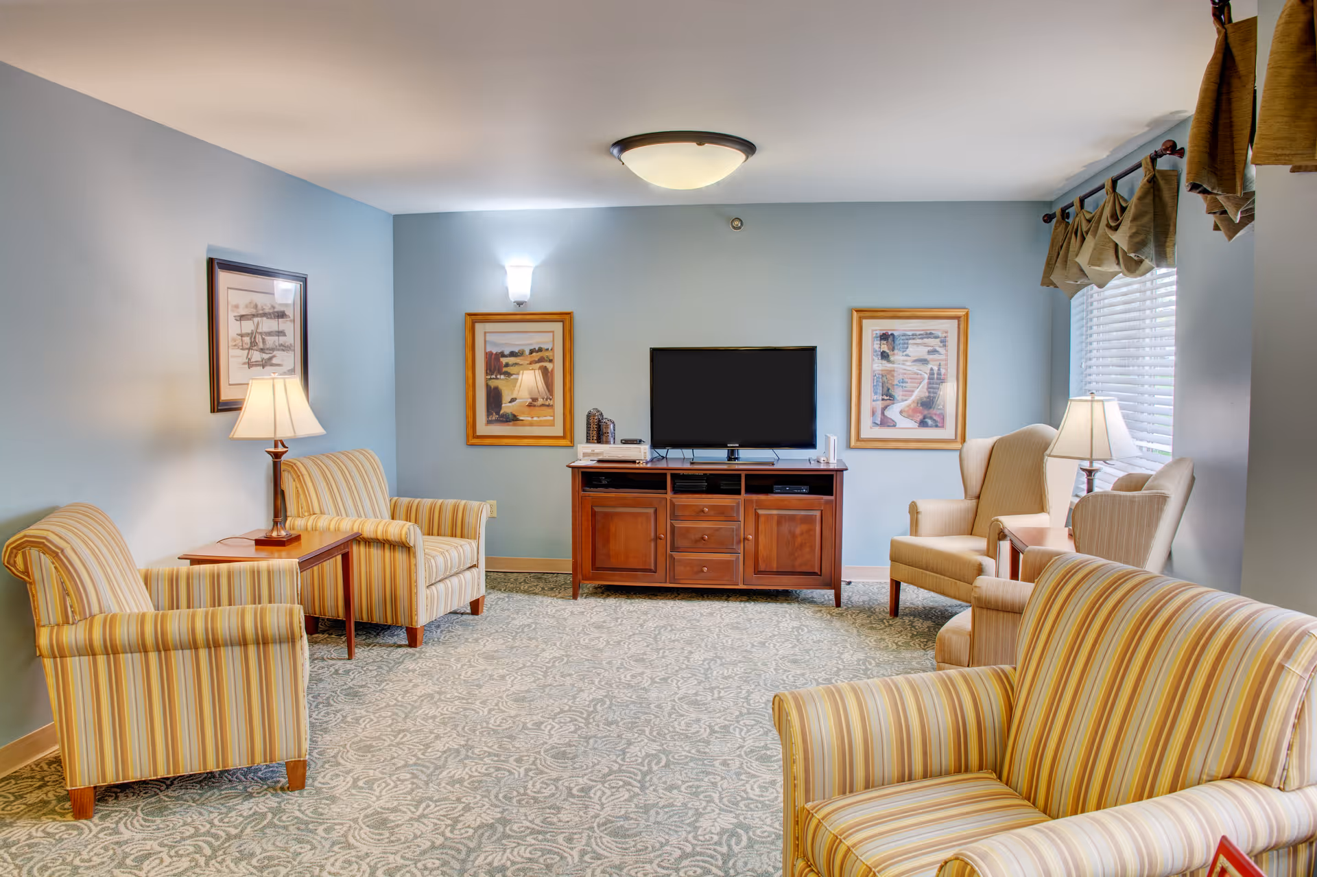 A cozy living room with light blue walls, patterned carpet, and several upholstered armchairs with striped and solid fabric. A wooden TV stand with a flat-screen TV is centered on the far wall, flanked by two framed paintings. There are table lamps on side tables next to the chairs, and a window with curtains on the right side.