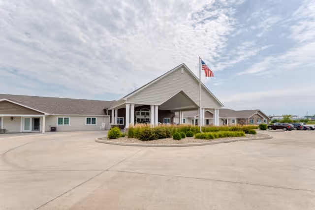 Exterior view of a single-story senior living facility building with a covered entrance supported by white columns. There is a landscaped roundabout with green shrubs and an American flag on a flagpole in front of the entrance. Several cars are parked to the right side of the building under a partly cloudy sky.
