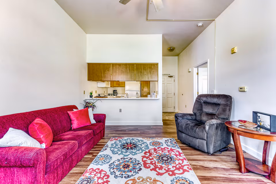 A bright living room with a red sofa adorned with red and white pillows on the left, a gray recliner chair on the right, and a colorful patterned rug on the wooden floor. Behind the sofa is a kitchen area with wooden cabinets and a white countertop. There is a small wooden side table next to the recliner with decorative items on it. The walls are white, and there is a ceiling fan above.