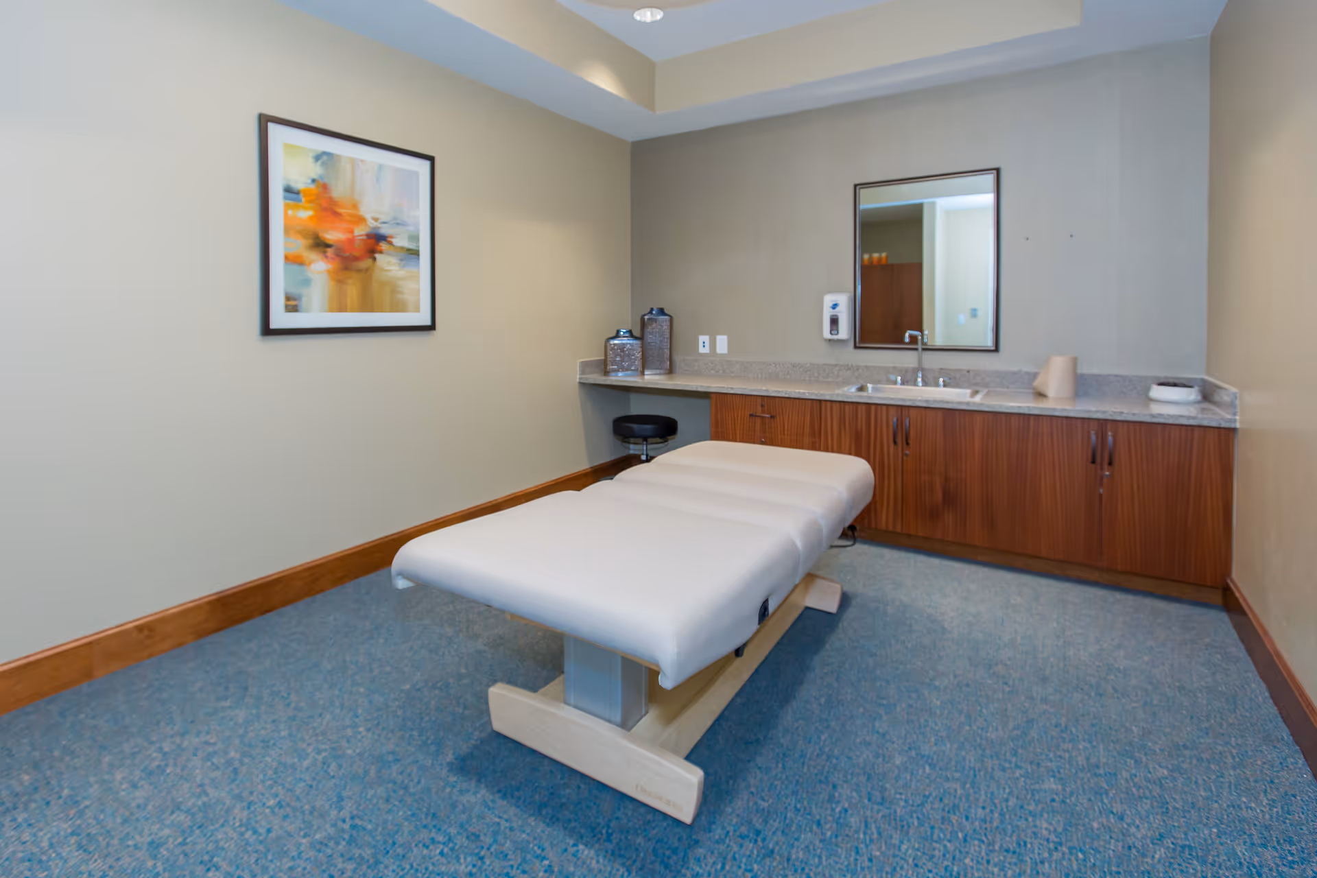 A therapy or massage room with a padded treatment table in the center, a countertop with a sink and cabinets along the back wall, a mirror above the sink, a stool tucked under the counter, and a colorful abstract painting on the left wall.