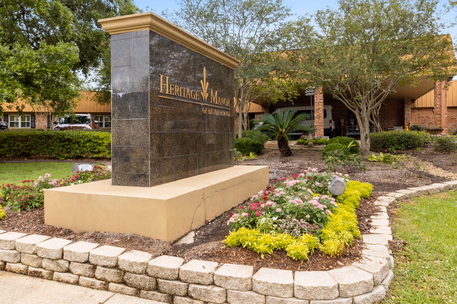 Outdoor view of Heritage Manor of Baton Rouge sign made of dark stone with gold lettering, surrounded by landscaped flower beds and greenery, with part of the building and trees in the background.