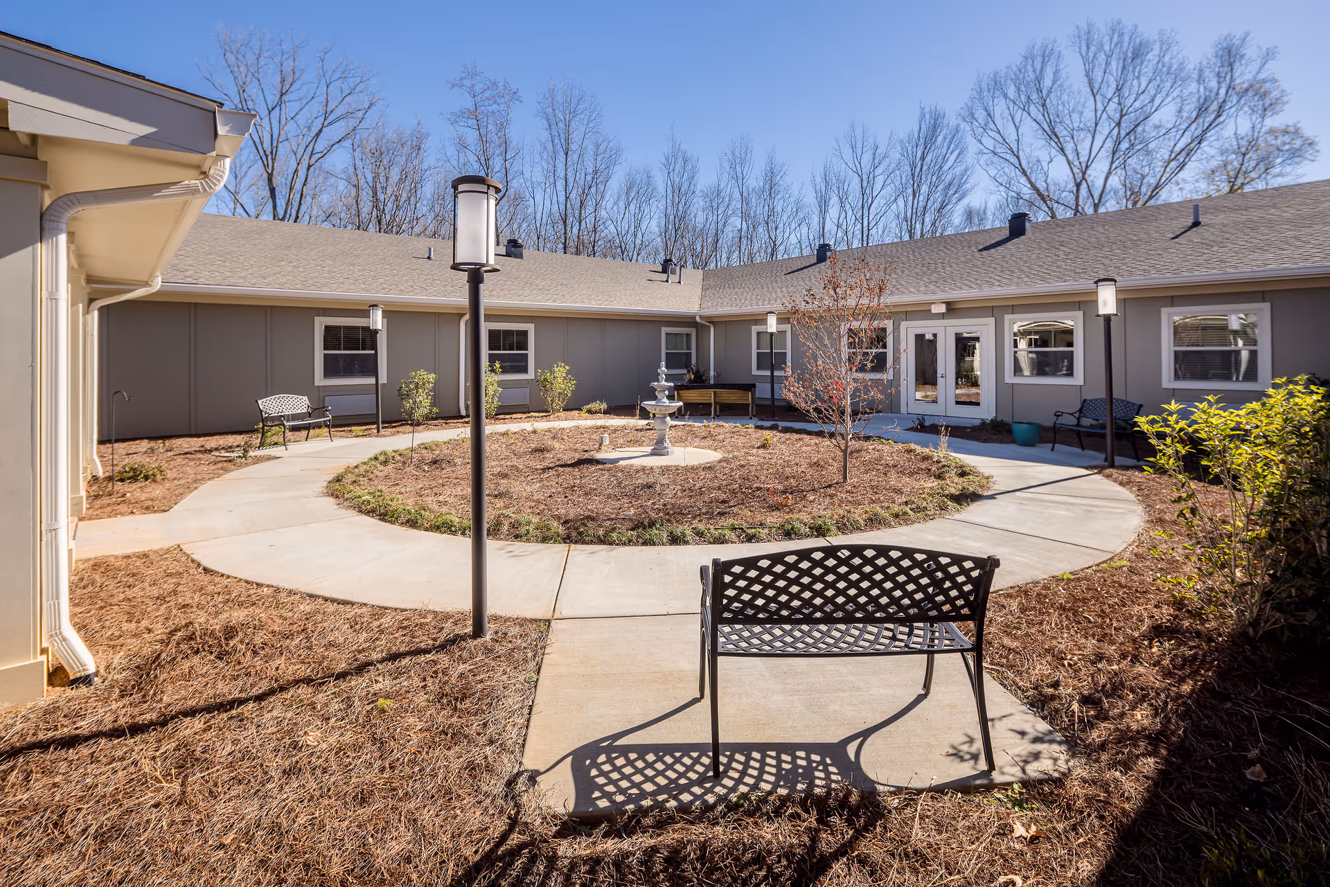 Outdoor courtyard area at Oaks at Eagle's Landing featuring a circular concrete walkway surrounding a mulched garden bed with a small fountain and young trees. Several black metal benches and lamp posts are placed along the walkway. The courtyard is enclosed by a single-story building with multiple windows and a double glass door entrance. Leafless trees are visible in the background under a clear blue sky.