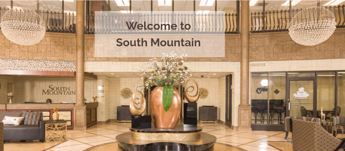 Interior view of the South Mountain Healthcare and Rehabilitation Center lobby featuring a large decorative vase with flowers on a round table in the center, two large chandeliers hanging from the ceiling, a reception desk on the left with the South Mountain sign behind it, and seating areas with sofas and chairs on both sides.
