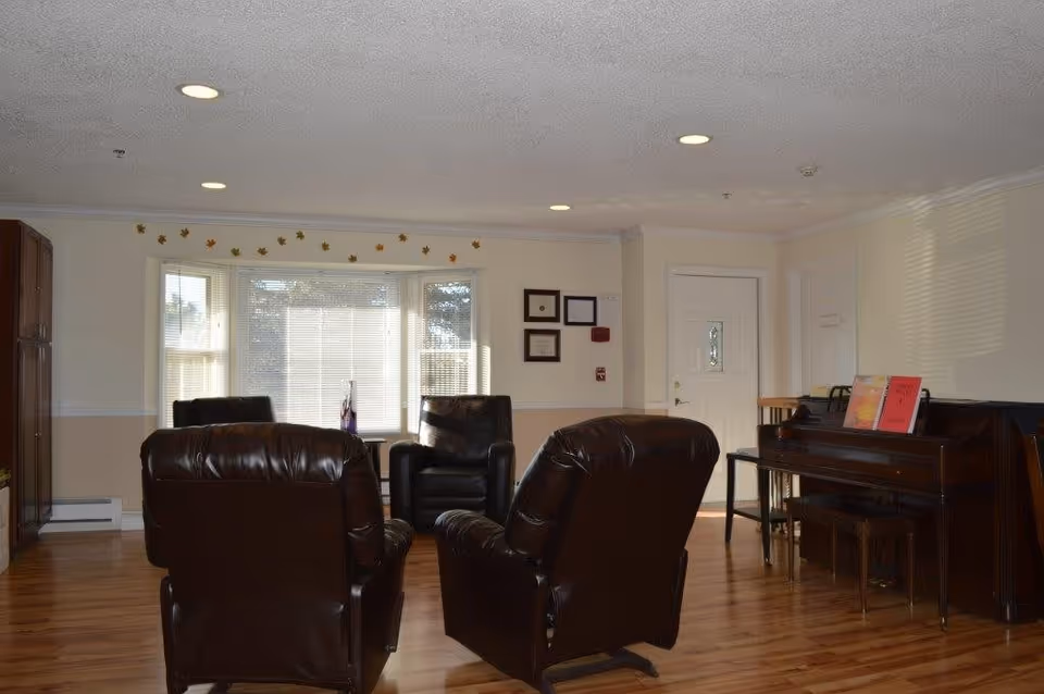 A cozy living room with four dark brown leather armchairs arranged around a central area. The room has light-colored walls, a large window with blinds, and a wooden floor. On the right side, there is a piano with sheet music on the stand, and a small table and chair near a white door. The walls have some framed certificates and a fire alarm.