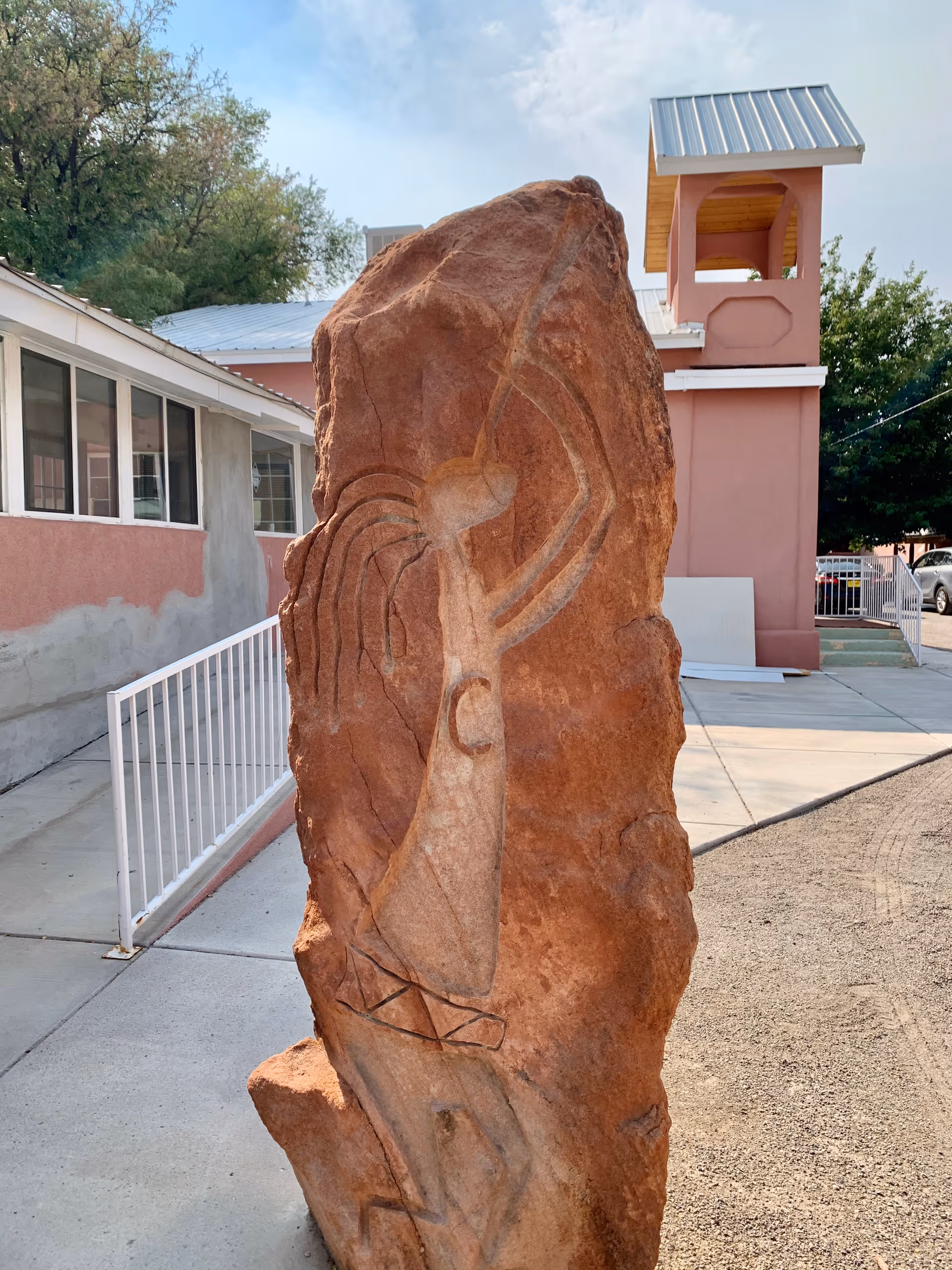 A tall reddish-brown stone sculpture with an abstract carved figure stands outdoors on a concrete pathway next to a pink building with white railings and windows. Trees and a blue sky are visible in the background.