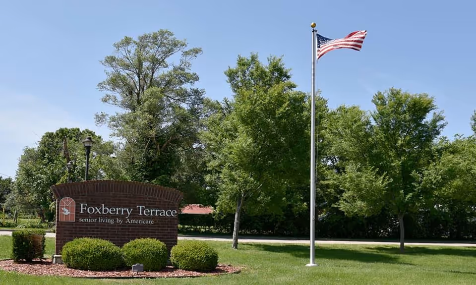 A brick sign for Foxberry Terrace senior living by Americare is displayed on a well-maintained lawn with green bushes and trees. An American flag on a tall flagpole is flying to the right of the sign under a clear blue sky.