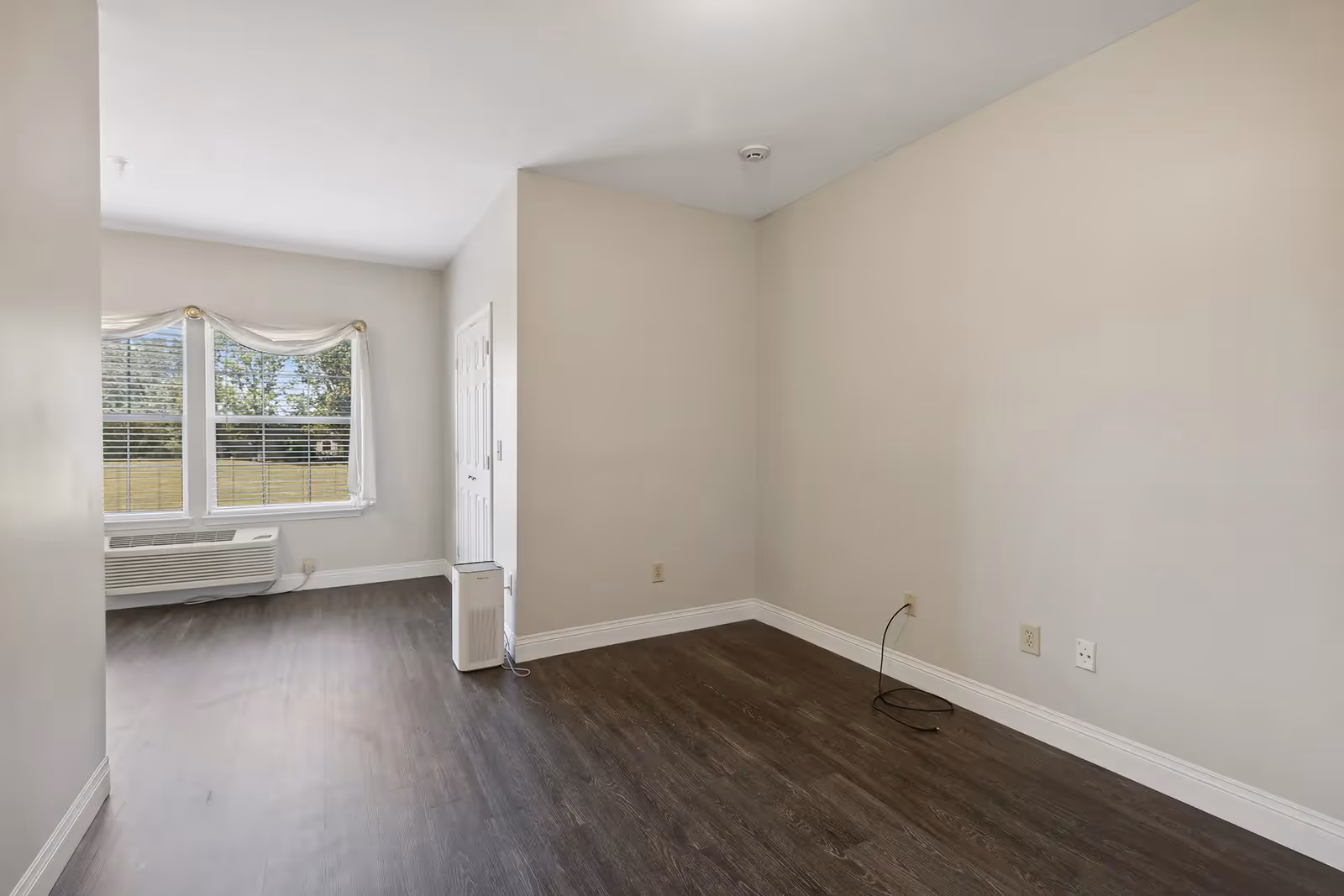 Empty room with light beige walls and dark wood flooring. There is a large window with white blinds and a sheer curtain, an air conditioning unit below the window, and a small white appliance or device on the floor near the corner. Electrical outlets and a cable are visible on the right wall.
