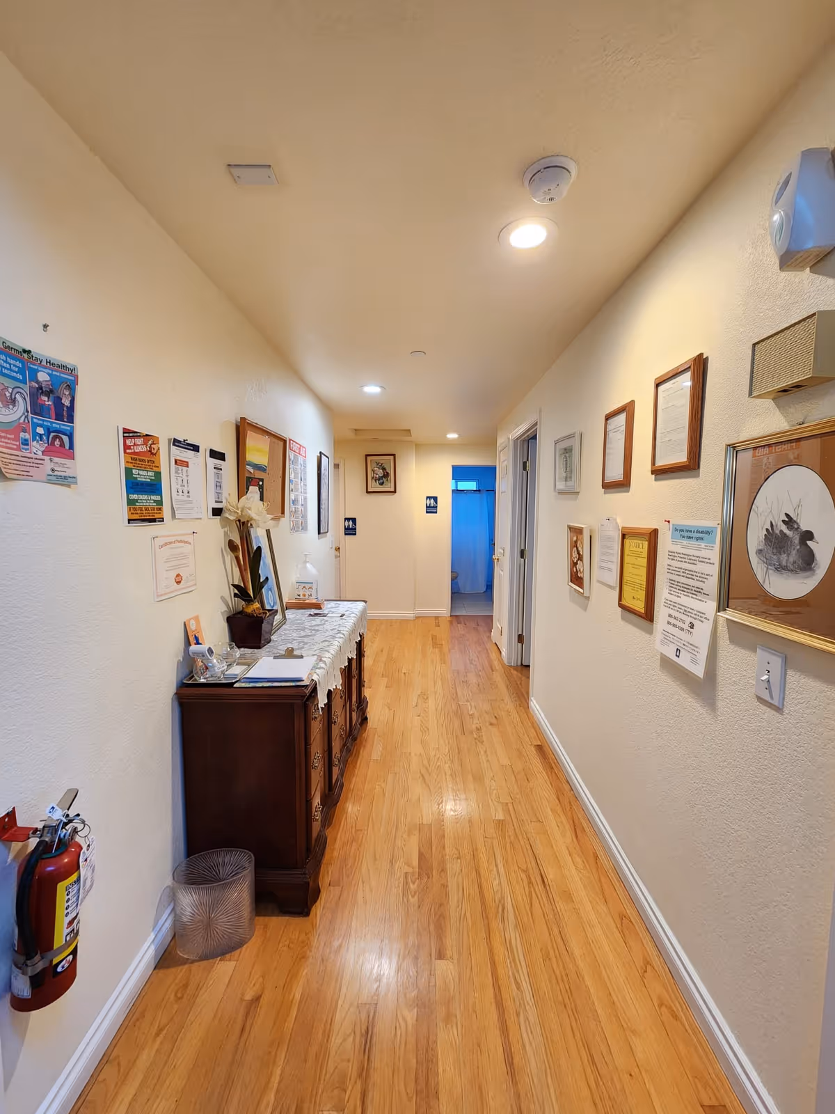 A well-lit hallway with wooden flooring and cream-colored walls. On the left side, there is a wooden cabinet with a white lace runner, a potted plant, and various papers and items on top. The walls on both sides are decorated with framed pictures, certificates, and informational posters. At the end of the hallway, there are two doors with restroom signs visible, and one door is slightly open showing a blue curtain inside. A fire extinguisher is mounted on the left wall near the foreground.