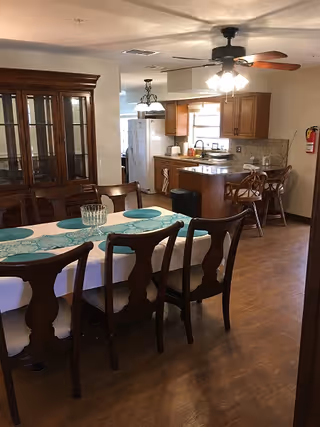 Interior view of a dining area and kitchen in an assisted living facility. The dining area features a wooden table with six chairs, a white tablecloth, and a blue patterned table runner with matching placemats. Behind the dining table is a large wooden china cabinet. The kitchen has wooden cabinets, a countertop with two bar stools, a ceiling fan with lights, and various kitchen appliances including a refrigerator and microwave. A fire extinguisher is mounted on the wall near the kitchen.