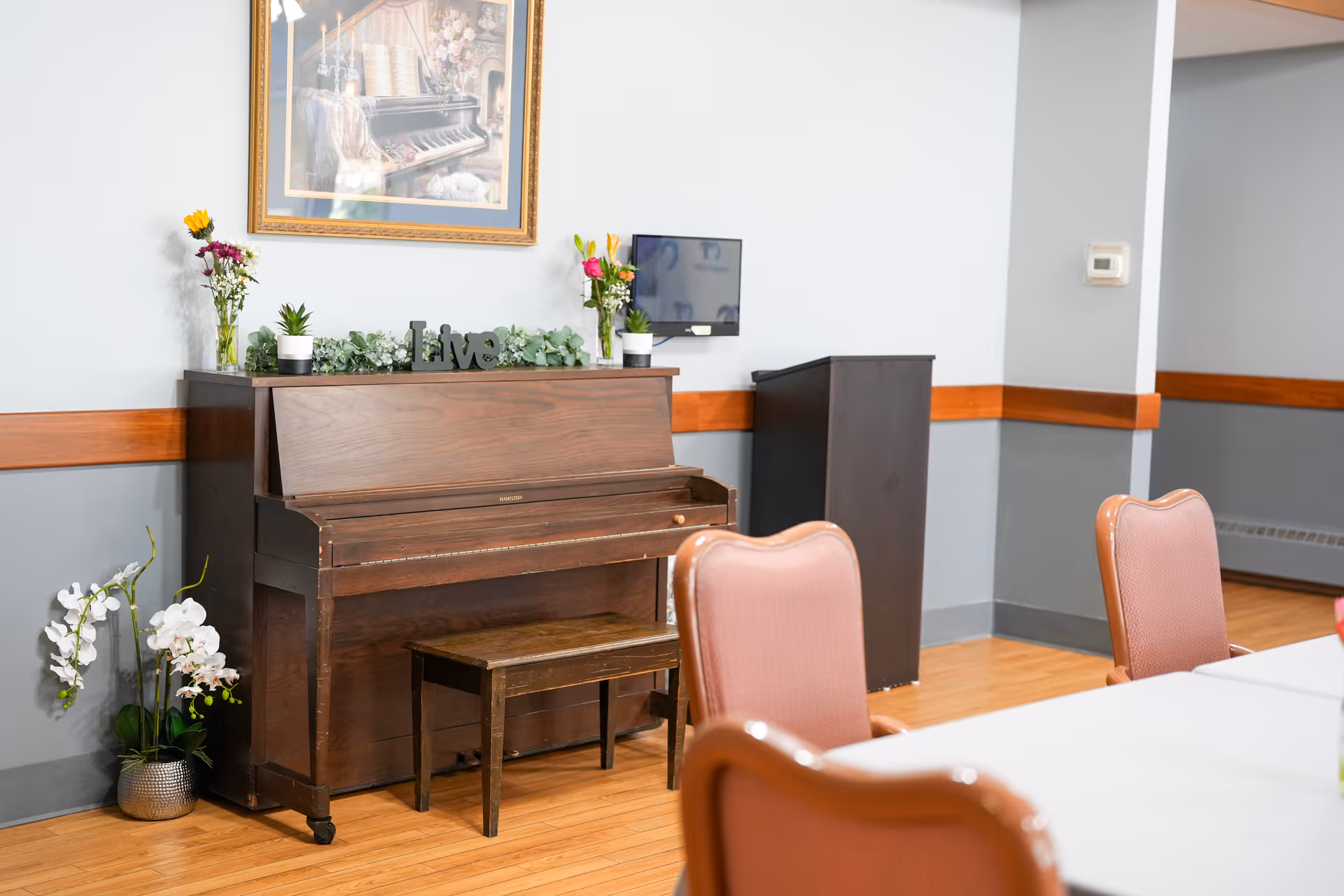 A room with a wooden upright piano and matching bench against a gray wall with a wooden chair rail. On top of the piano are two vases with flowers, small potted plants, and a decorative 'live' sign. Above the piano hangs a framed picture of a piano with flowers. To the right of the piano is a small black podium and a wall-mounted thermostat. In the foreground are pink cushioned chairs and a white table.
