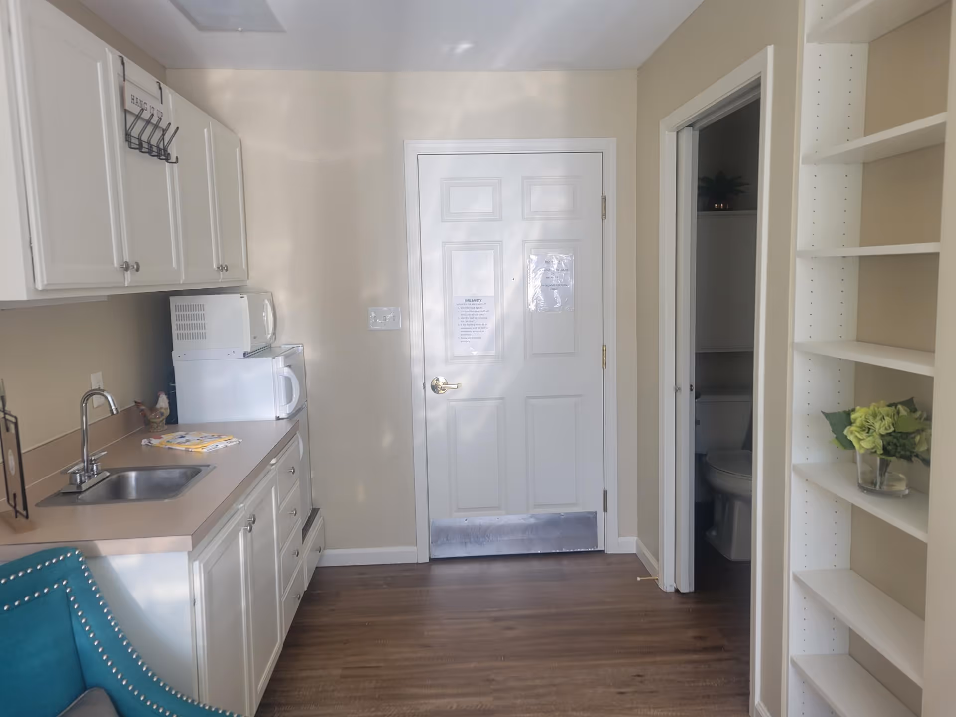 Small kitchenette with white cabinets, sink, microwave and fridge, a door in the center, shelving on the right and a glimpse of a bathroom.