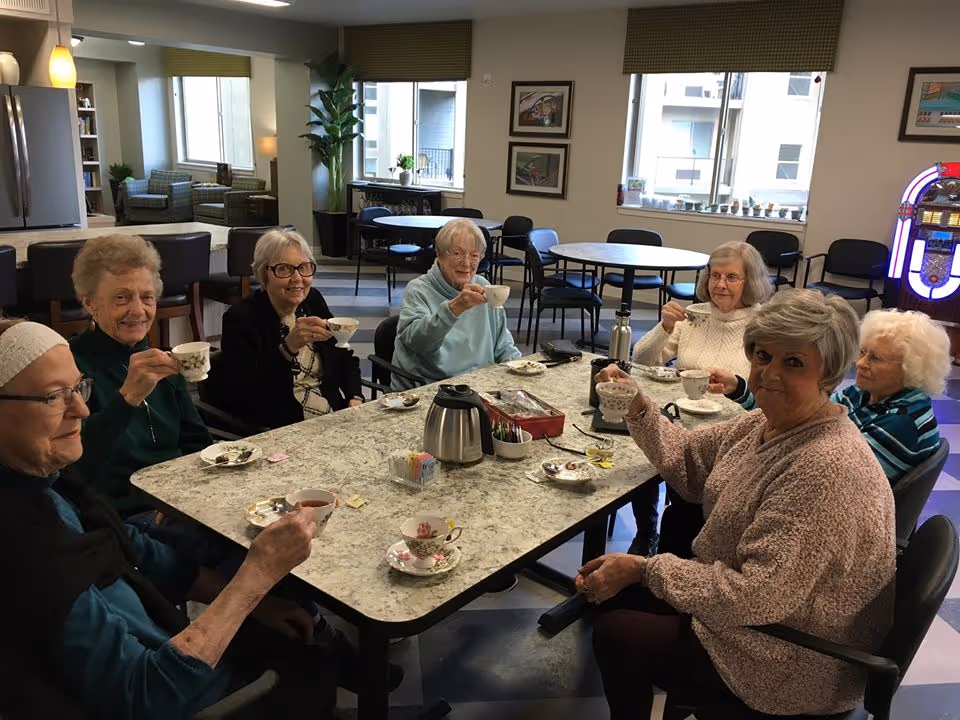Seven elderly women sitting around a rectangular table in a well-lit room, holding teacups and smiling. The table has a coffee pot, sugar packets, and small plates. The room has large windows, framed pictures on the wall, and a jukebox in the background.
