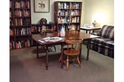 A cozy room with a wooden table and chair in the center, surrounded by bookshelves filled with books. There is a plaid sofa on the right side and a small side table with a lamp. The room has carpeted flooring and framed artwork on the wall.