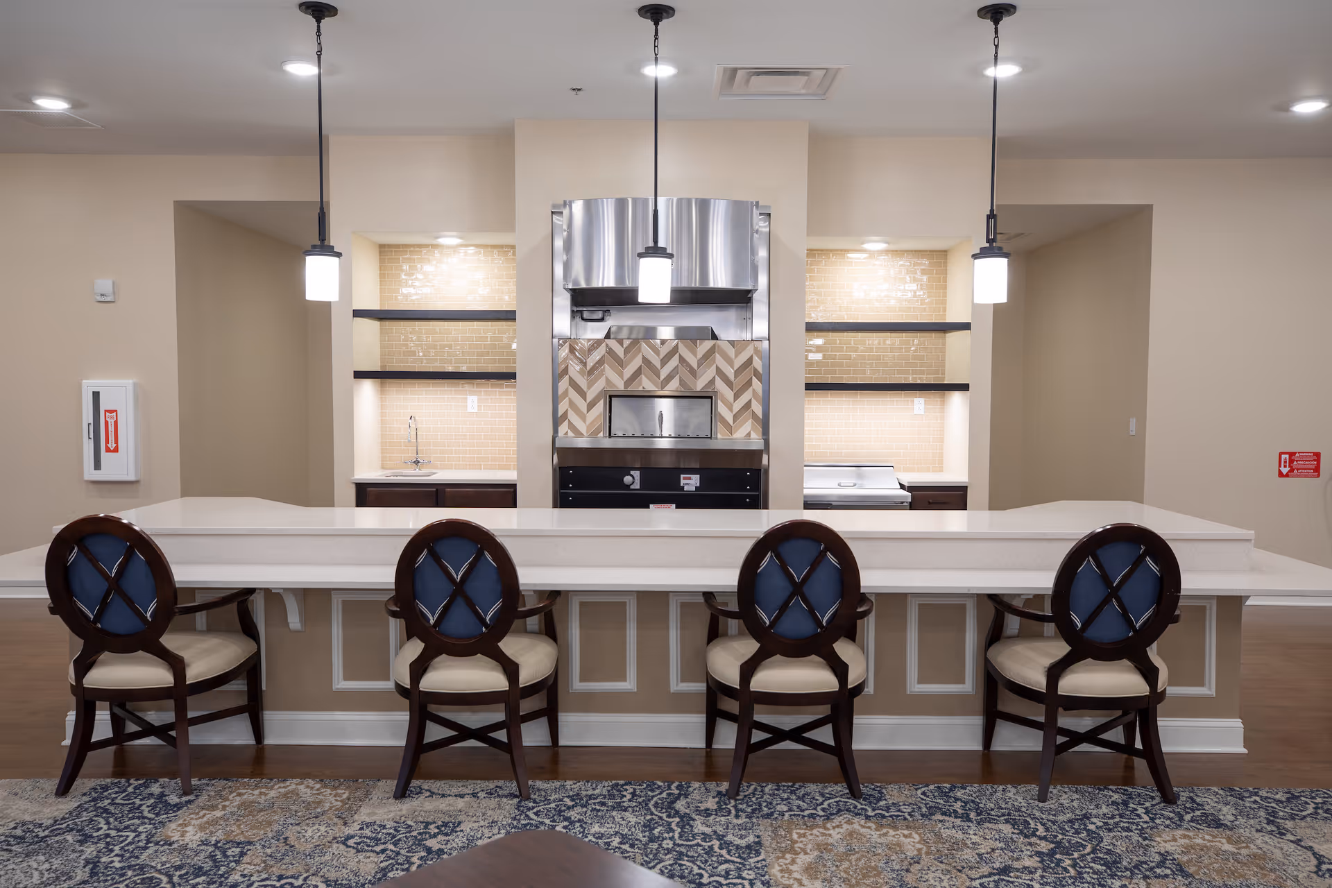 A modern kitchen area with a long white countertop and four wooden chairs with blue cushioned backs and beige seats. Behind the counter are built-in shelves with lighting, a stainless steel oven with a decorative tile backsplash, and two pendant lights hanging from the ceiling. The floor has a patterned rug in front of the counter.