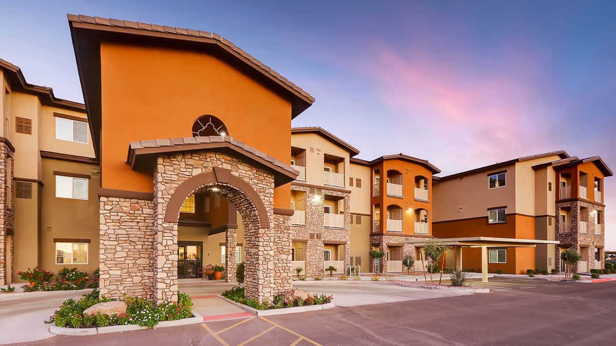 Front exterior of a three-story senior living building with a stone-arched entrance, balconies, and landscaped driveway at sunset.