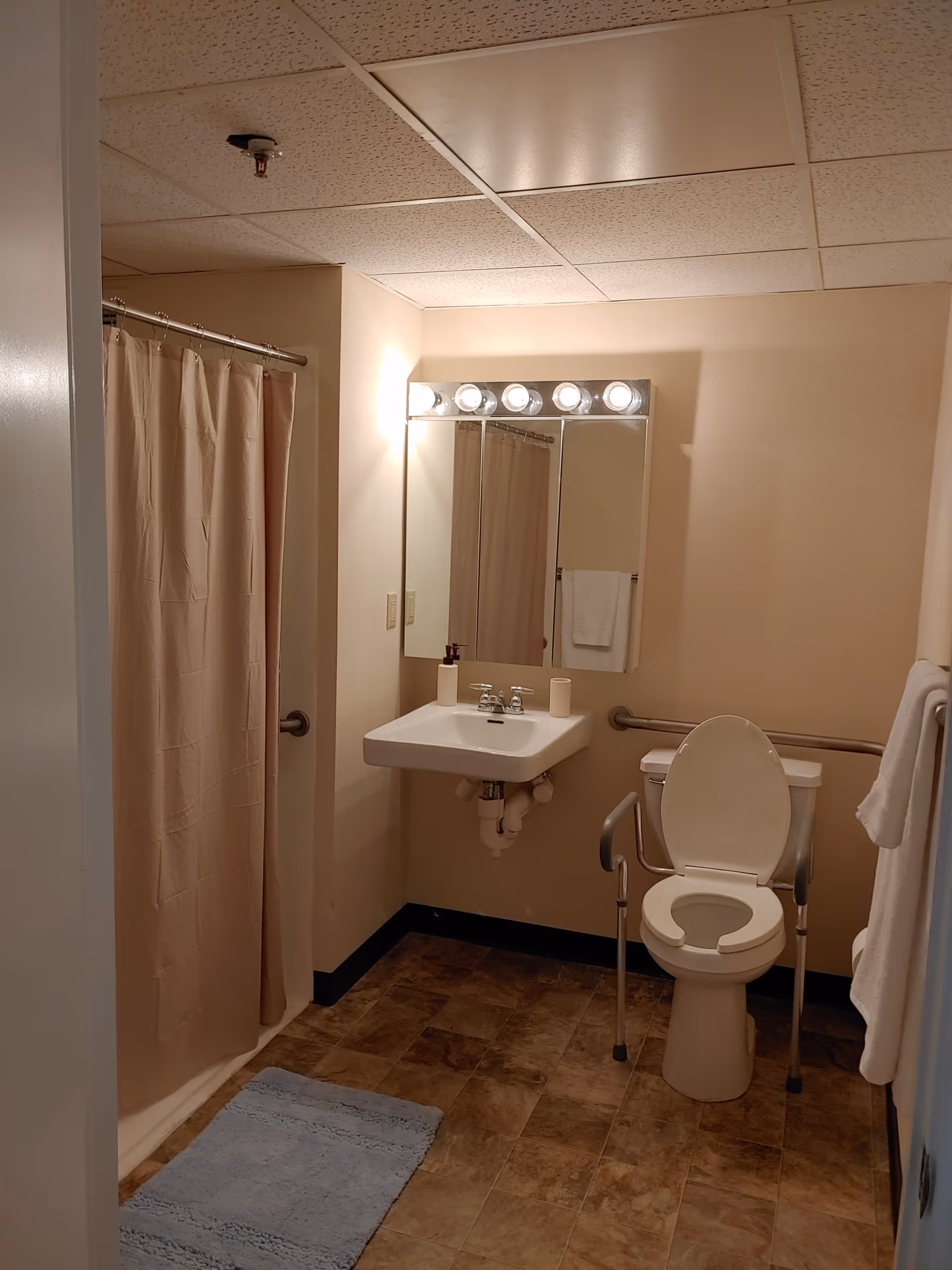 A bathroom with a beige shower curtain on the left, a white sink with a mirror and lights above it in the center, and a toilet with support rails on the right. There is a blue bath mat on the floor and a towel hanging on the wall next to the toilet.