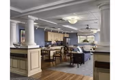 A spacious common area in a senior living facility featuring a kitchen with wooden cabinets and a countertop, a dining area with tables and chairs, and a living room section with sofas and armchairs. The room has decorative columns, recessed lighting, and a ceiling fan, with a mix of hardwood and carpeted flooring.