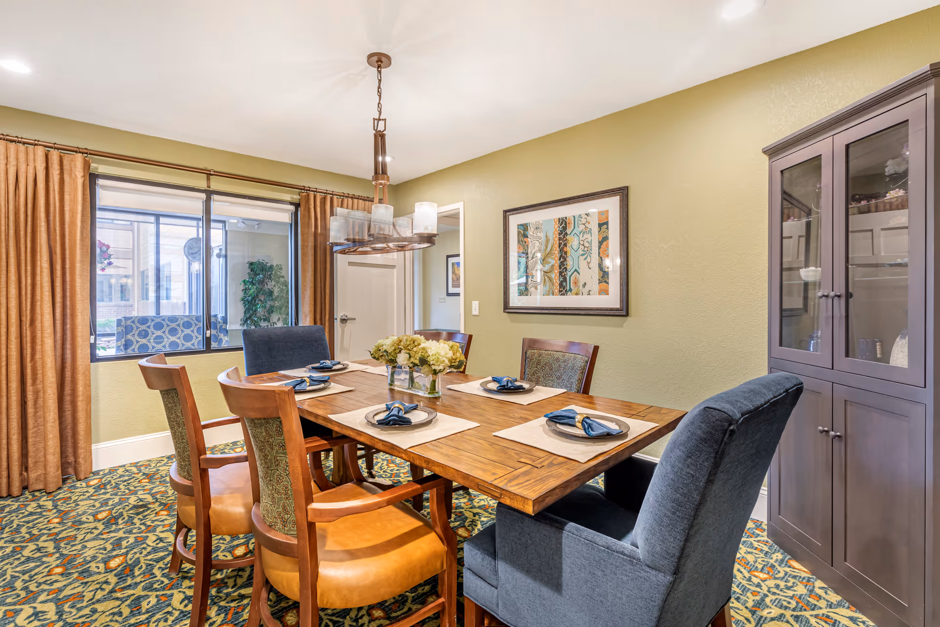 A dining room with a wooden table set for four, featuring blue napkins on white plates and a centerpiece of flowers. The room has patterned carpet, green walls, a framed artwork, a glass-front cabinet, and large windows with brown curtains.