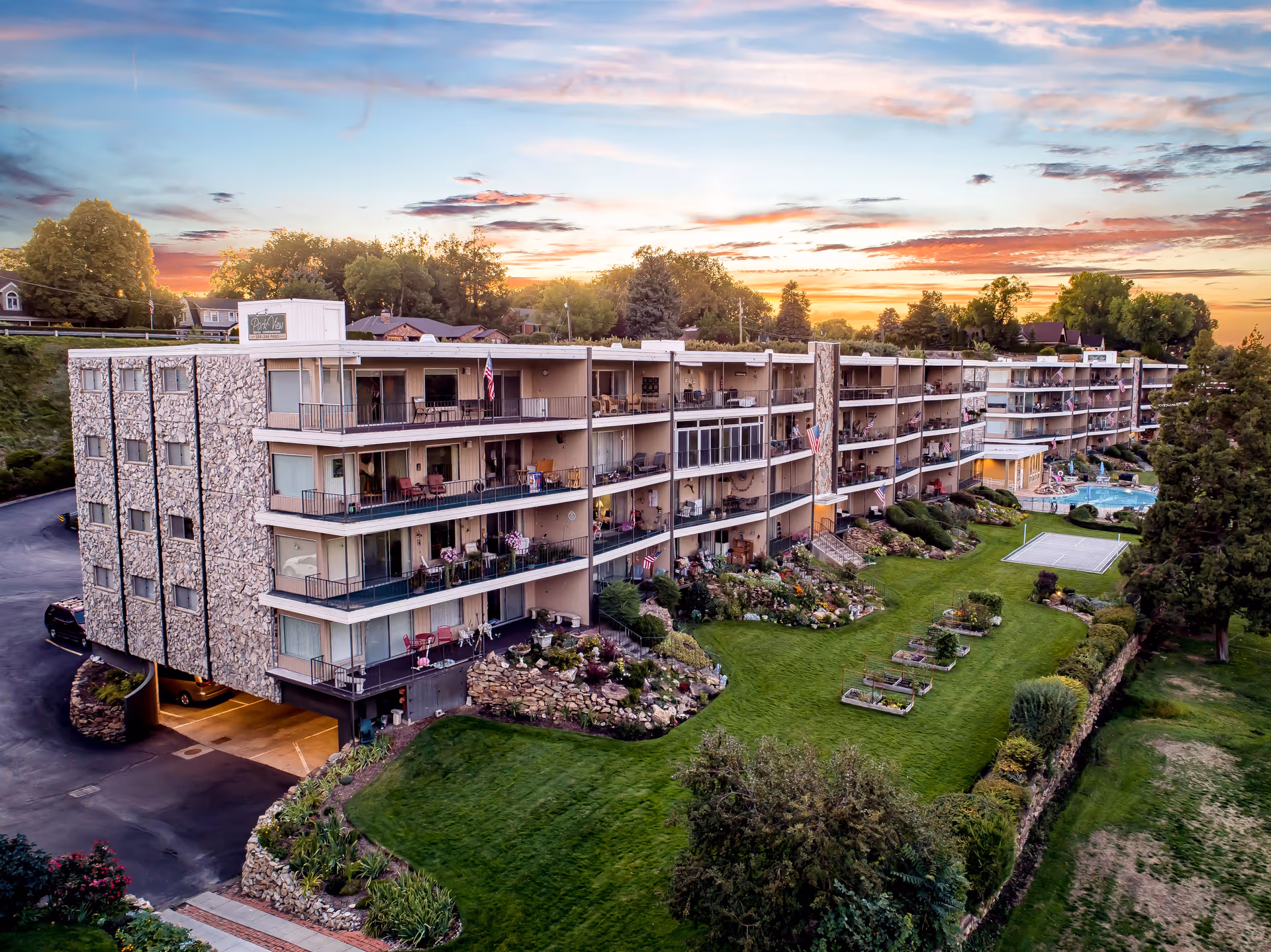 Multi-story senior living building with balconies, manicured lawn, and a pool under a sunset sky.