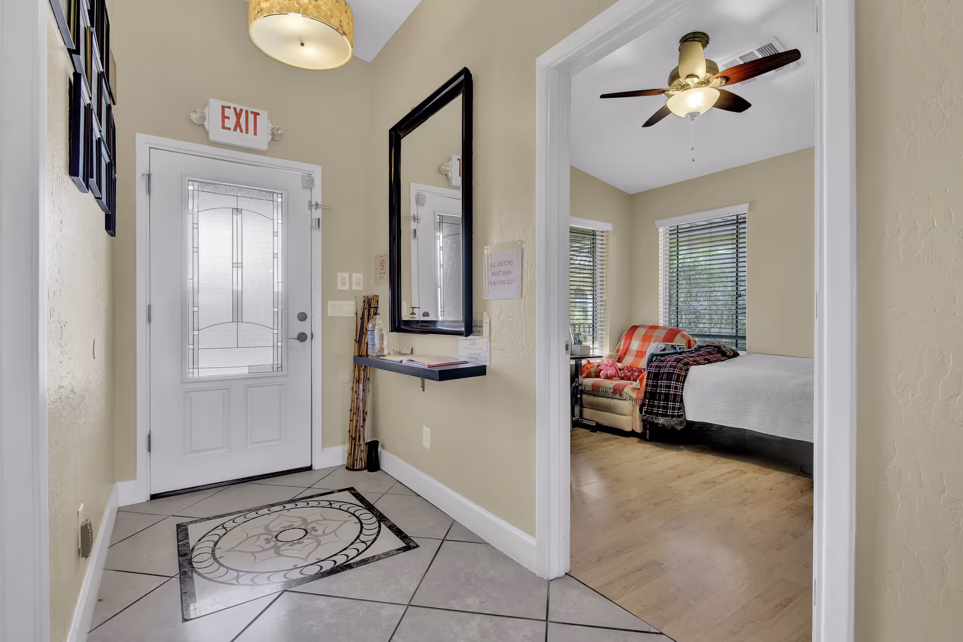 View of an entryway with a decorative tiled floor and a white door with frosted glass and an EXIT sign above it. To the right, there is a doorway leading to a bedroom with a bed, a ceiling fan, and a chair covered with a red and white checkered blanket. A large mirror hangs on the wall next to a small shelf with a hand sanitizer bottle and a visitor sign-in sheet.