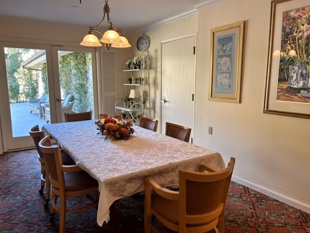 A dining room with a long rectangular table covered with a white patterned tablecloth and decorated with a centerpiece of autumn-themed flowers and foliage. There are eight wooden chairs with brown cushions around the table. The room has a patterned carpet, cream-colored walls with framed artwork, a clock, and a shelving unit with decorative items. A glass door opens to an outdoor patio area with greenery.