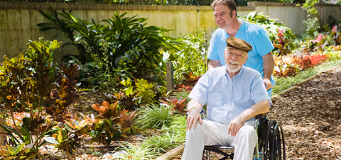 A caregiver pushes an elderly man in a wheelchair along a leafy outdoor garden path.