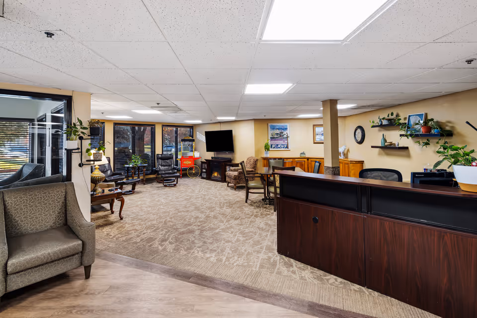Spacious senior living community lobby with a reception desk, seating area, TV, and decorative plants.