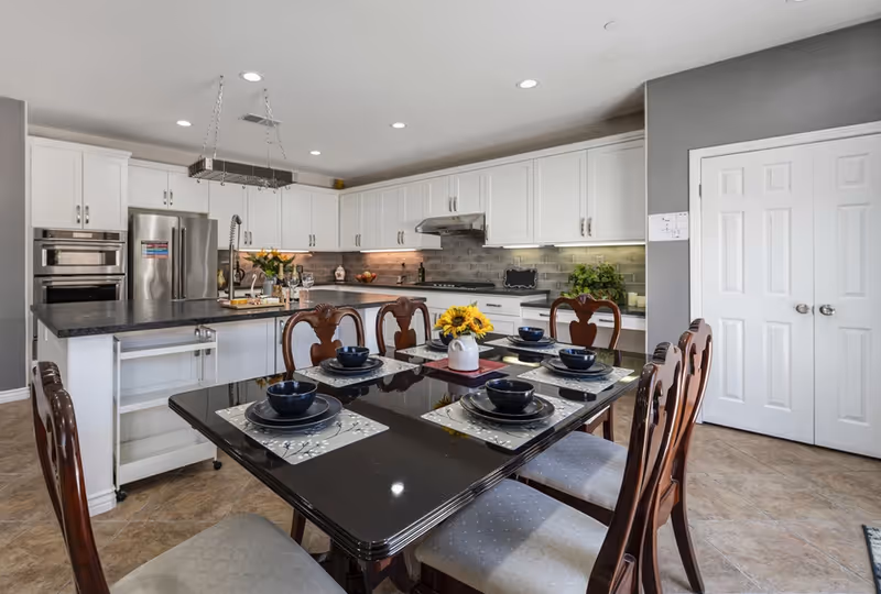 A modern kitchen and dining area with white cabinets, stainless steel appliances, and a black dining table set for six with dark blue bowls and plates. The kitchen has a large island with a sink and a hanging rack above it. The floor is tiled, and there are double white doors on the right side of the room.