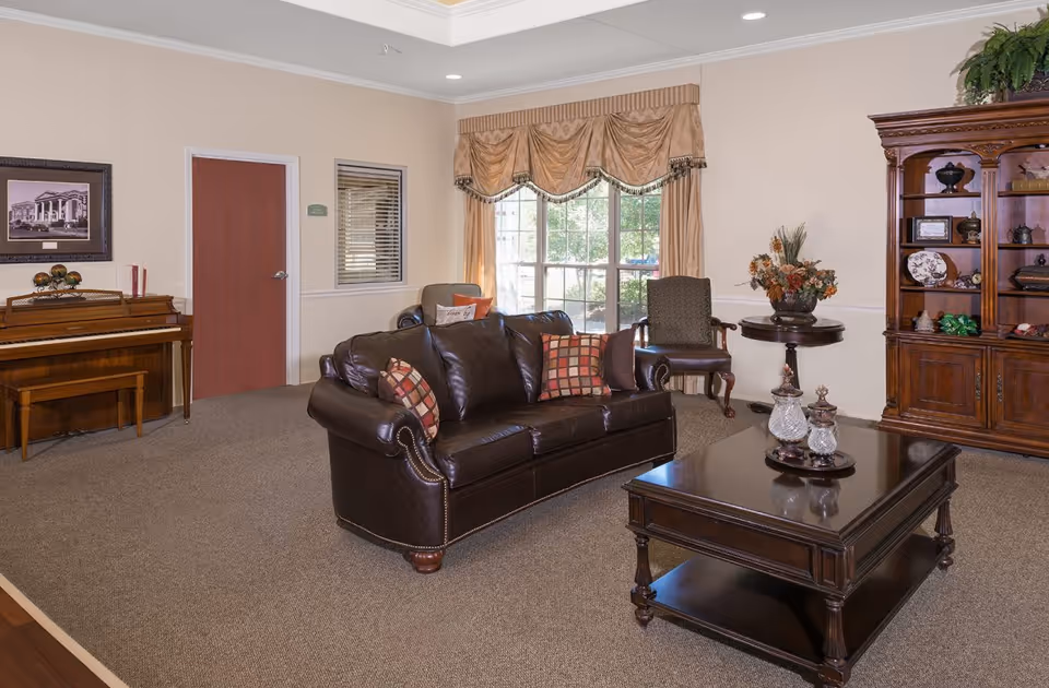 A cozy living room with a dark brown leather sofa adorned with patterned pillows, a wooden coffee table with decorative items, a wooden cabinet filled with ornaments, a piano with a bench, and a window with beige curtains letting in natural light.