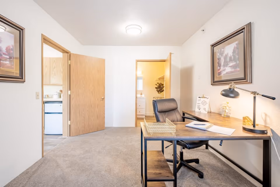 A bright room with beige carpet featuring a wooden desk with a black office chair. On the desk are a black desk lamp, a clipboard with paper, a notebook, and a wire paper tray. Two framed landscape paintings hang on the white walls. Two open wooden doors reveal a kitchen area with a refrigerator and a bathroom with a vanity and a plant.