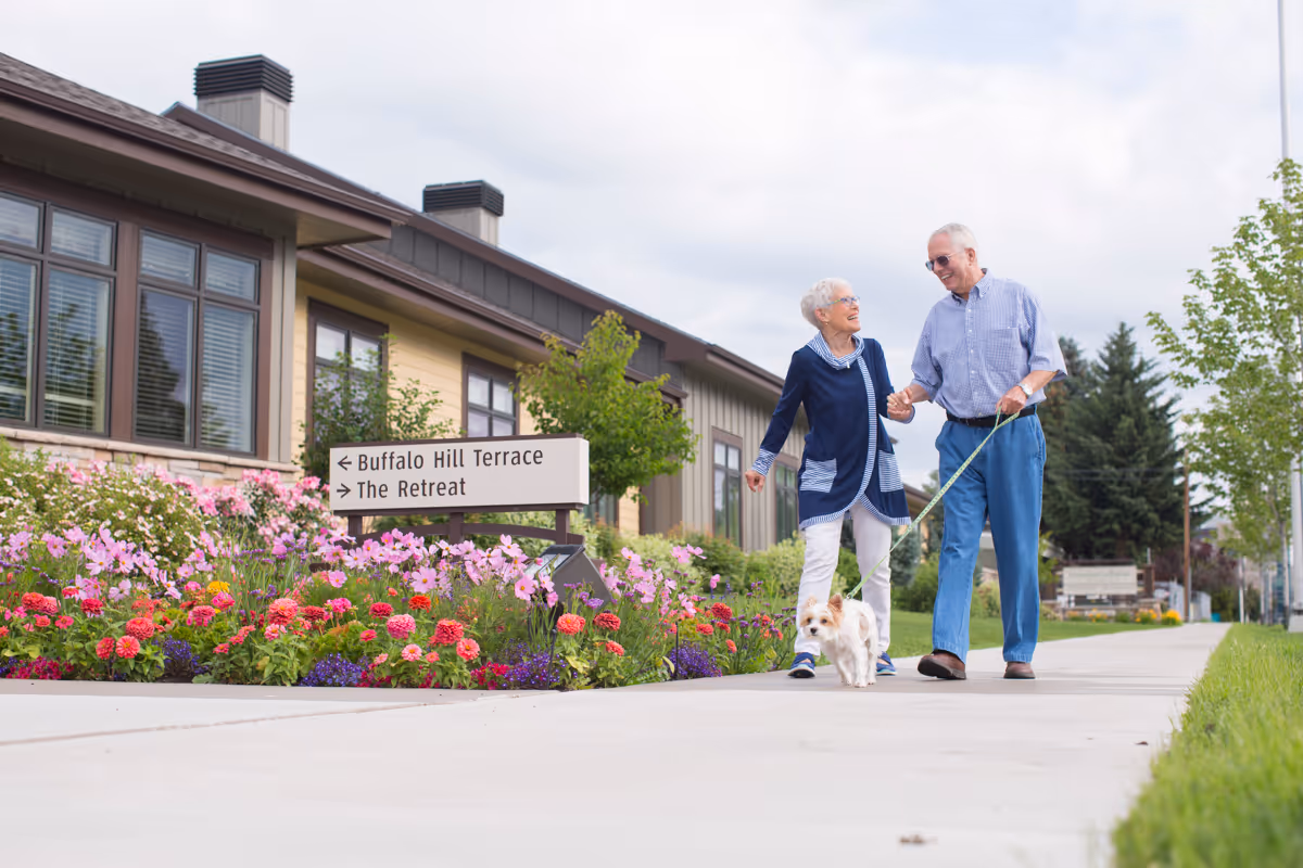 An elderly couple walking hand in hand on a sidewalk outside a building, with the man holding a small dog on a leash. There are colorful flowers and greenery along the side of the building, and a sign pointing to Buffalo Hill Terrace and The Retreat.