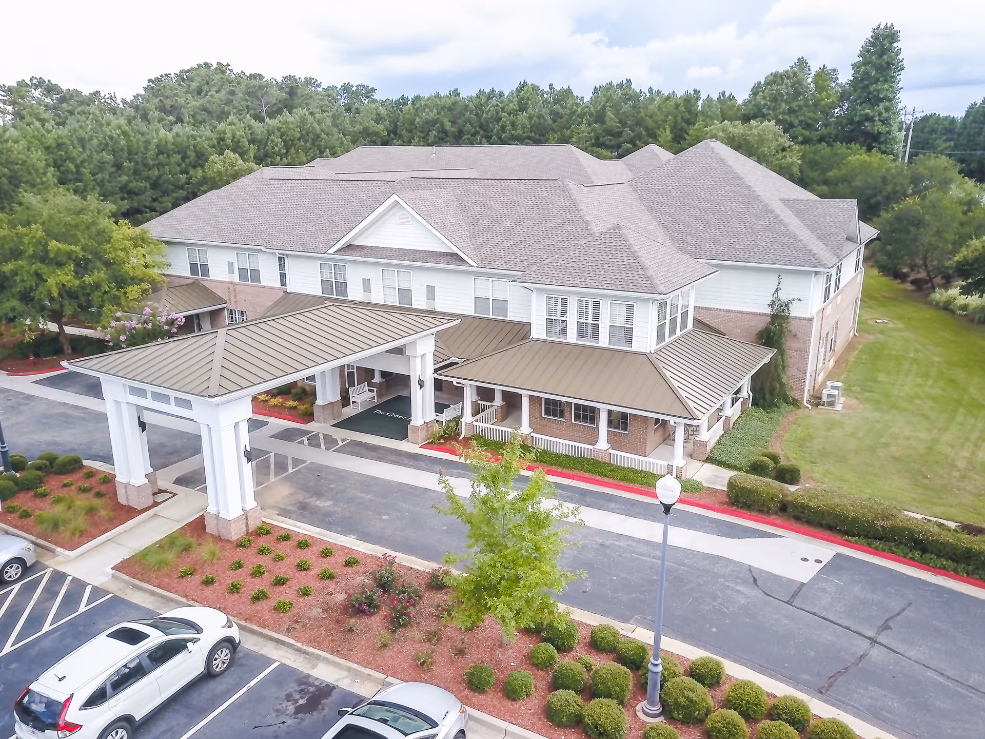 Aerial view of a two-story senior living building with a covered entrance porte-cochere, parking lot, and landscaped grounds.