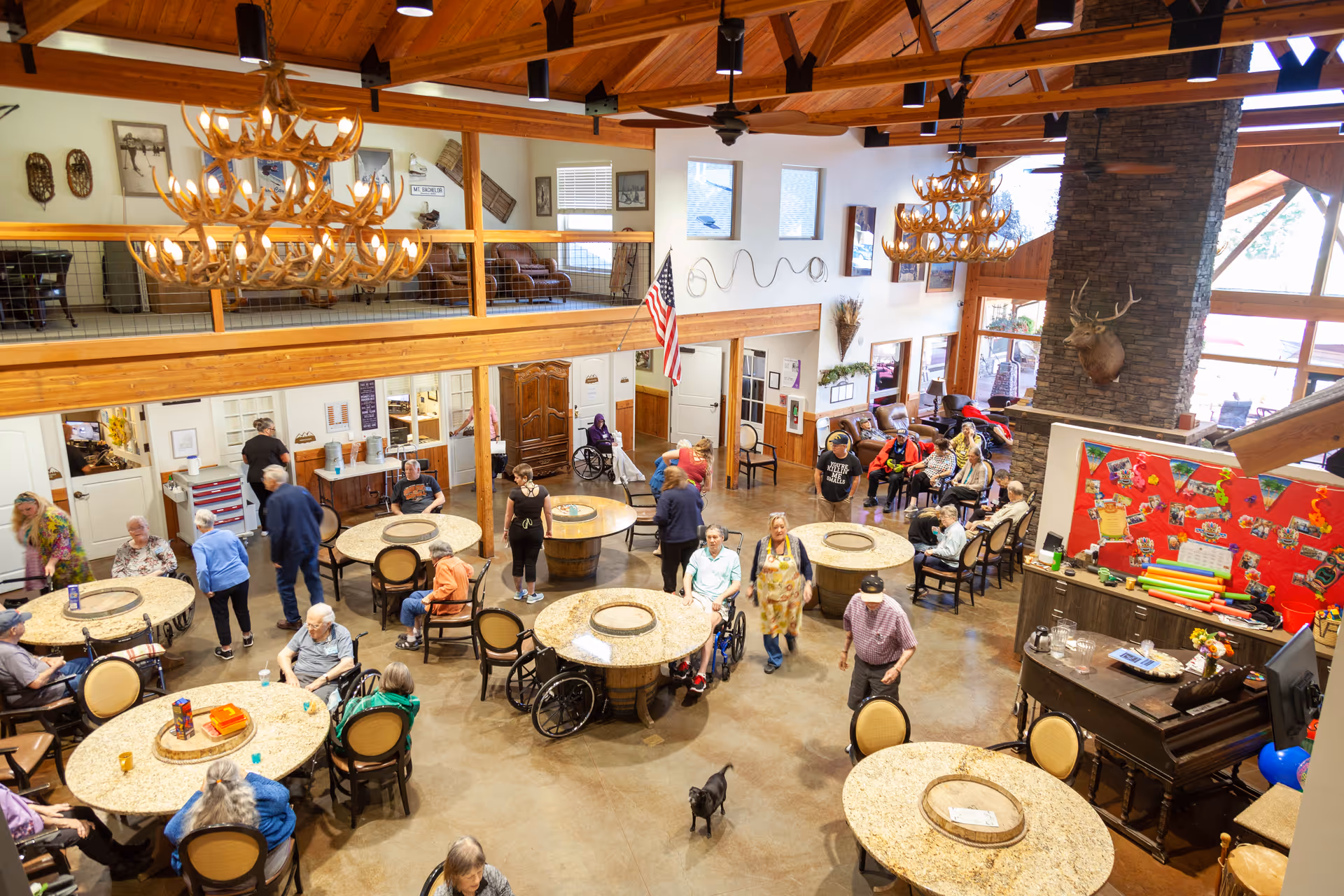 A spacious, well-lit common area in a senior living facility with multiple round tables and chairs. Several elderly residents, some in wheelchairs, are seated or standing around the room. The room features high wooden ceilings with large antler chandeliers, a stone fireplace with a mounted deer head, and an American flag. There is a red bulletin board with decorations and various items on a counter near the fireplace. The atmosphere appears lively and social.