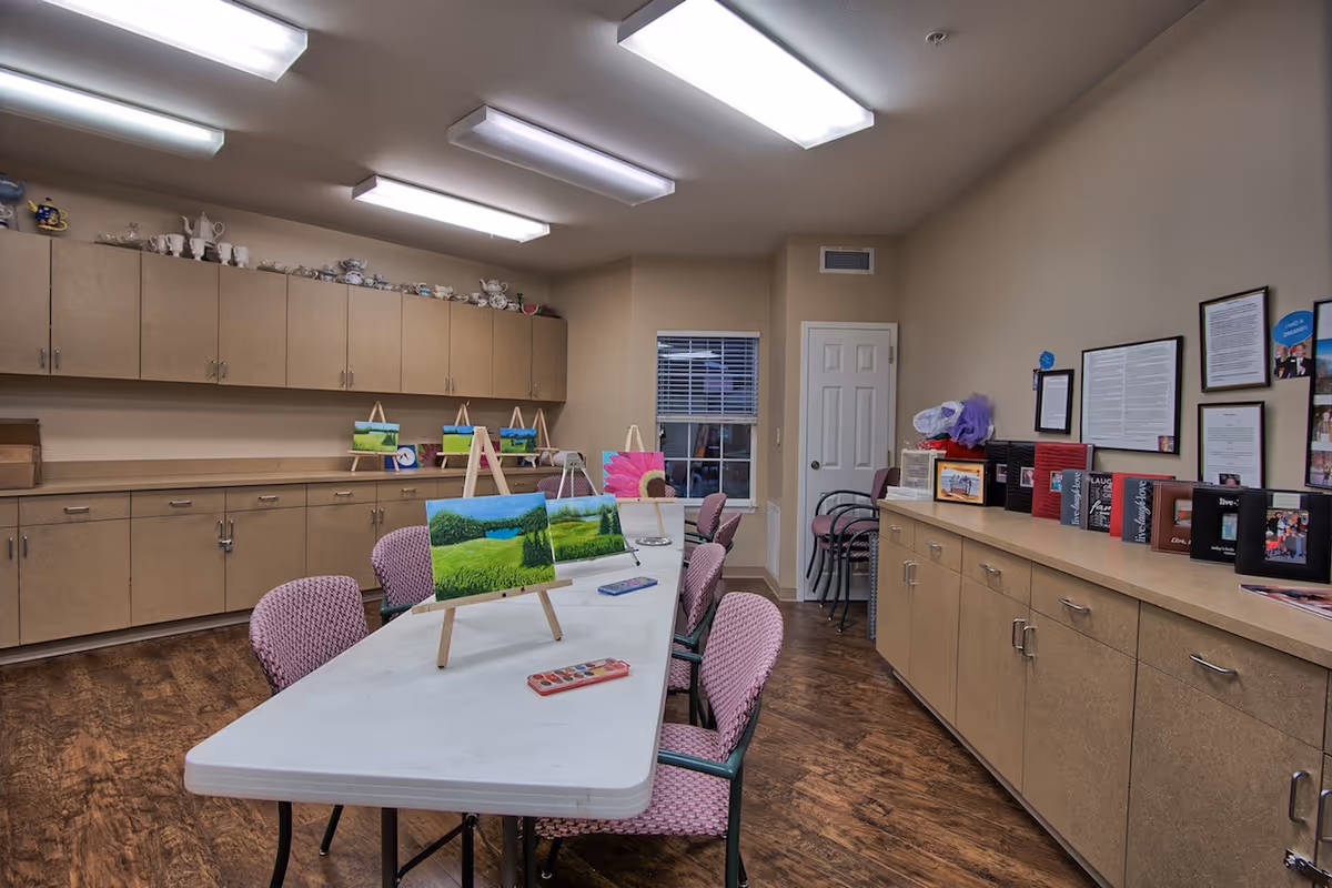 Well-lit activity room with tables and chairs, easels displaying small landscape paintings, and storage cabinets lining the walls.
