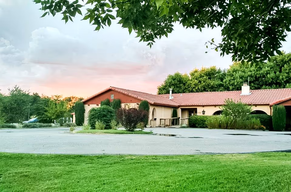 Exterior view of a single-story senior living facility building with a red tiled roof, surrounded by greenery and trees, under a partly cloudy sky at sunset.