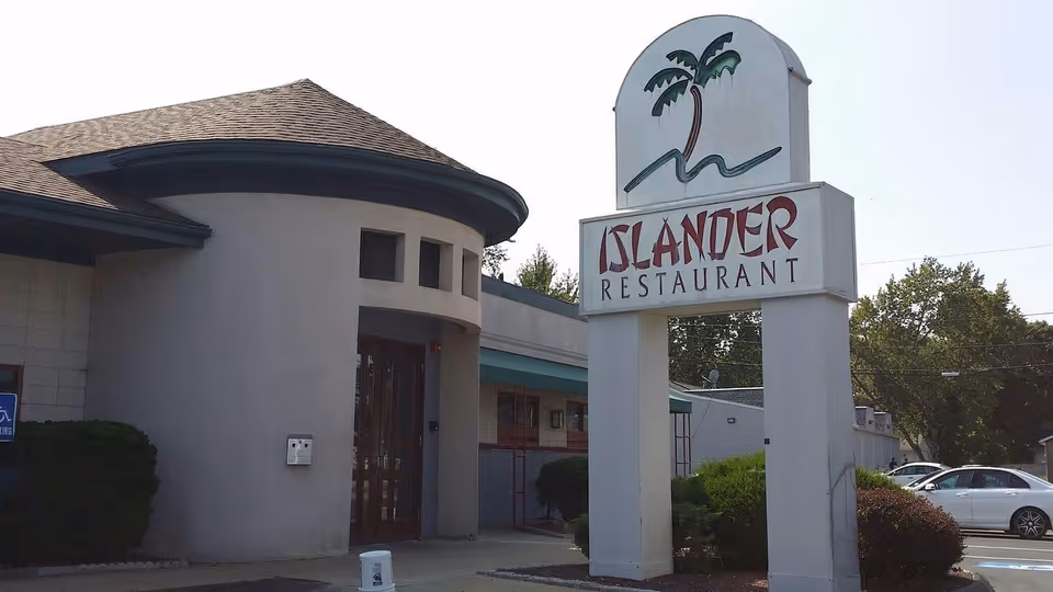Exterior view of a building with a rounded entrance and a large sign that reads 'Islander Restaurant' featuring a palm tree graphic. There are bushes and parked cars visible in the background under a clear sky.