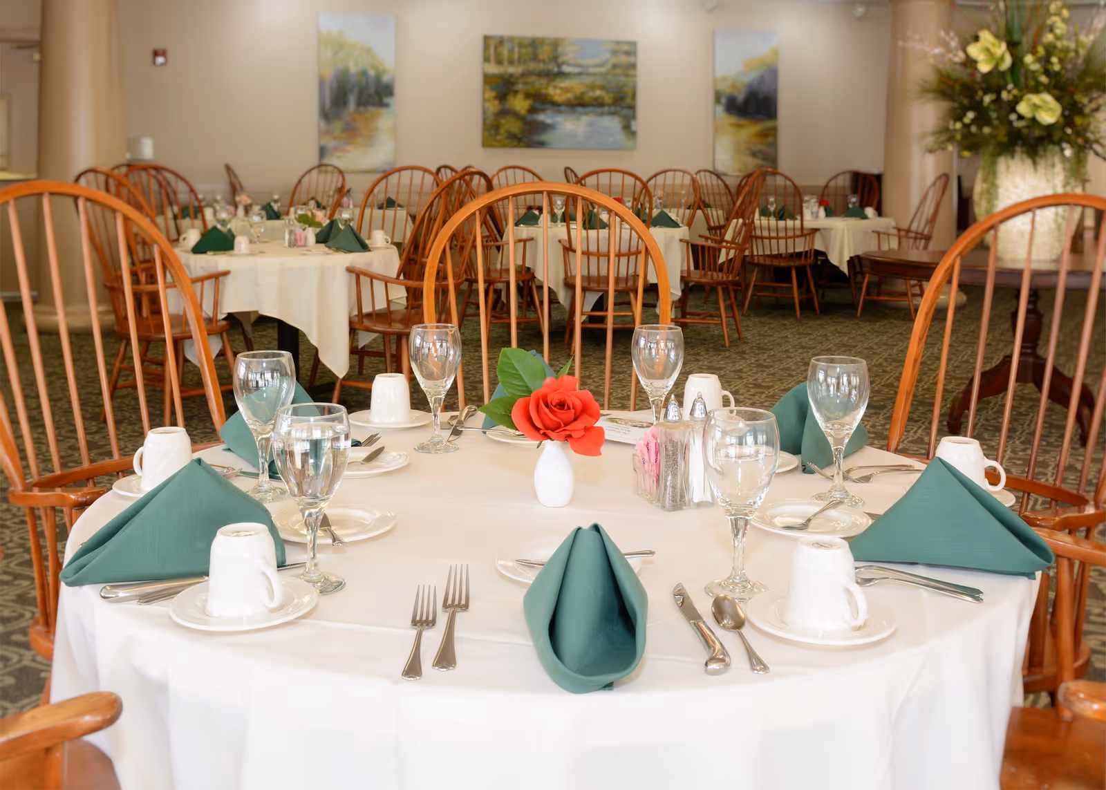 A dining room with round tables covered in white tablecloths, each set with green folded napkins, glassware, white cups, silverware, and a small vase with a red flower. Wooden chairs surround the tables, and paintings hang on the beige walls in the background.