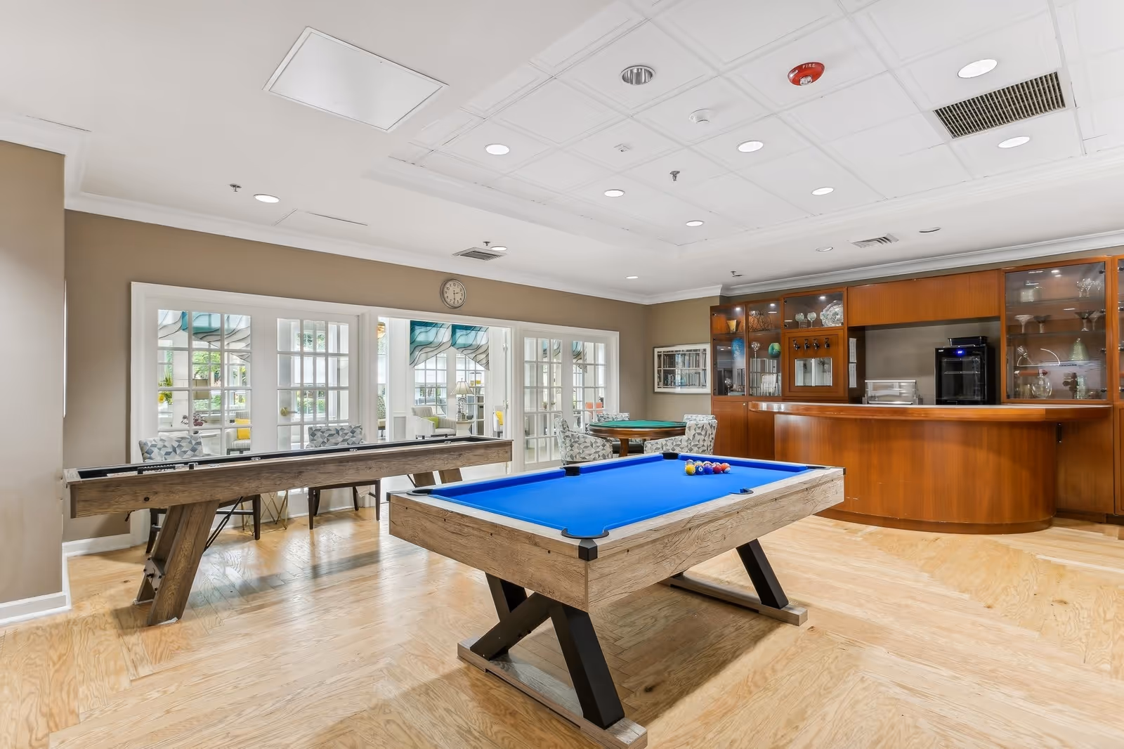 A bright recreational room featuring a blue felt pool table and a shuffleboard table on a wooden floor. The room has beige walls, a white ceiling with recessed lighting, and a wooden bar area with glass cabinets displaying glassware. There are several chairs and a small round table near large windows and glass doors that lead to an outdoor patio area.