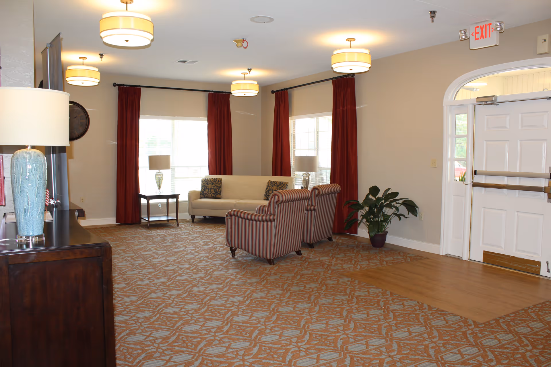 A spacious living room area with patterned carpet and beige walls. The room features a beige sofa with two patterned cushions, two striped armchairs, two side tables with lamps, red curtains on the windows, a large clock on the wall, a potted plant near the exit door, and multiple ceiling lights. The exit door is white with a glass panel and an illuminated red EXIT sign above it.