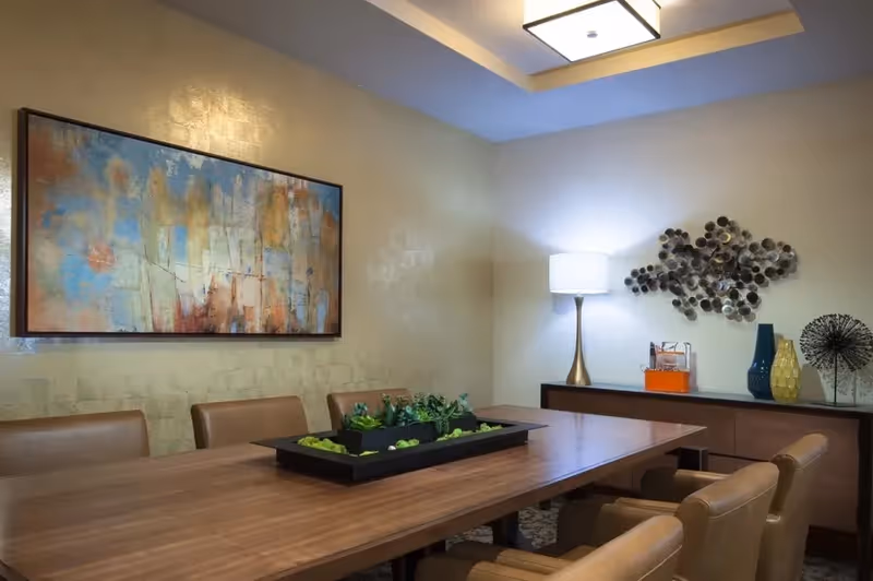 A modern dining room with a large wooden table surrounded by tan leather chairs. On the table is a rectangular planter with green plants. The walls are light-colored with abstract artwork and decorative wall sculptures. A table lamp with a white shade sits on a sideboard along with decorative vases and objects. The ceiling has a recessed light fixture.