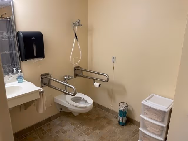 Accessible bathroom with a wall-mounted toilet equipped with two metal grab bars on either side. There is a white sink with a soap dispenser and a paper towel dispenser mounted on the wall. A shower curtain is partially visible on the left side. On the right side, there is a plastic storage drawer unit with folded towels and a small trash can. The walls are beige and the floor has small square tiles in neutral tones.